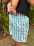 Wahine Holoholo Shorts 2.0 in ʻEhukai feature a Palaka pattern. Modeled outdoors with a child held on the hip, showcasing the relaxed fit.