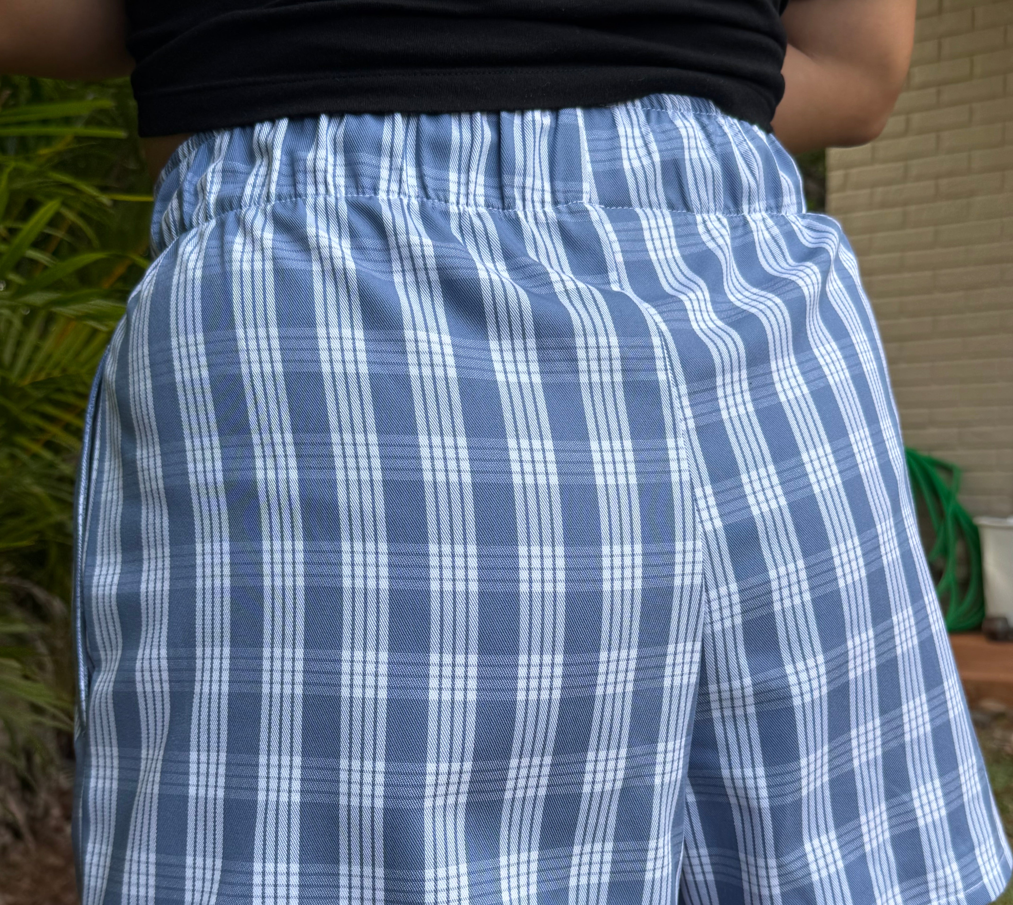 Wahine Holoholo Shorts 2.0 with a Palaka print, shown from the back. Elastic waistband, light blue tone, outdoors setting.