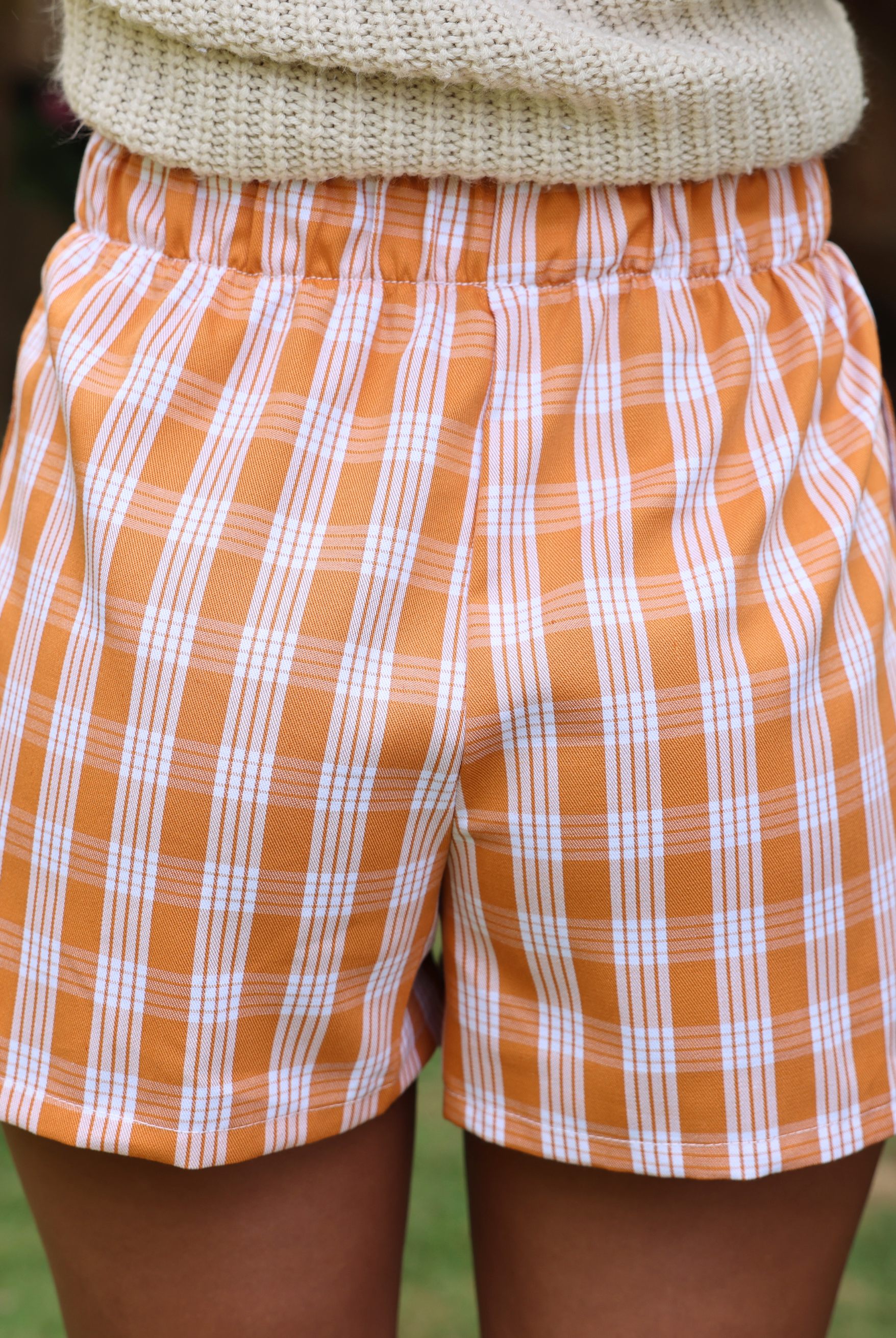 Wahine Holoholo Shorts 2.0 in Kaunaʻoa Rust Palaka print. Elastic waistband, paired with a beige knit top, outdoor setting.