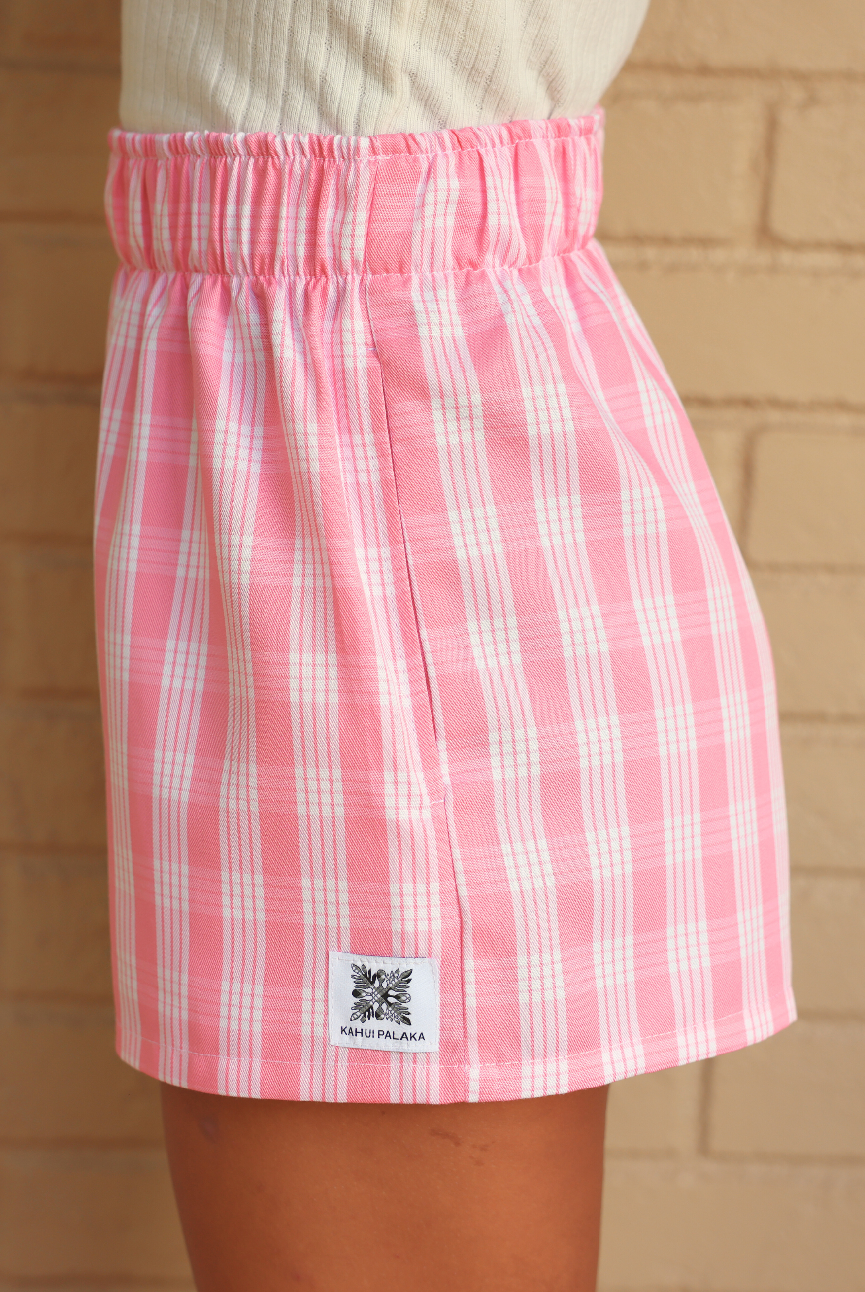 Wahine Holoholo Shorts 2.0 in Lokelani Light, featuring a pink Palaka print. Side view, elastic waistband, and logo tag visible.