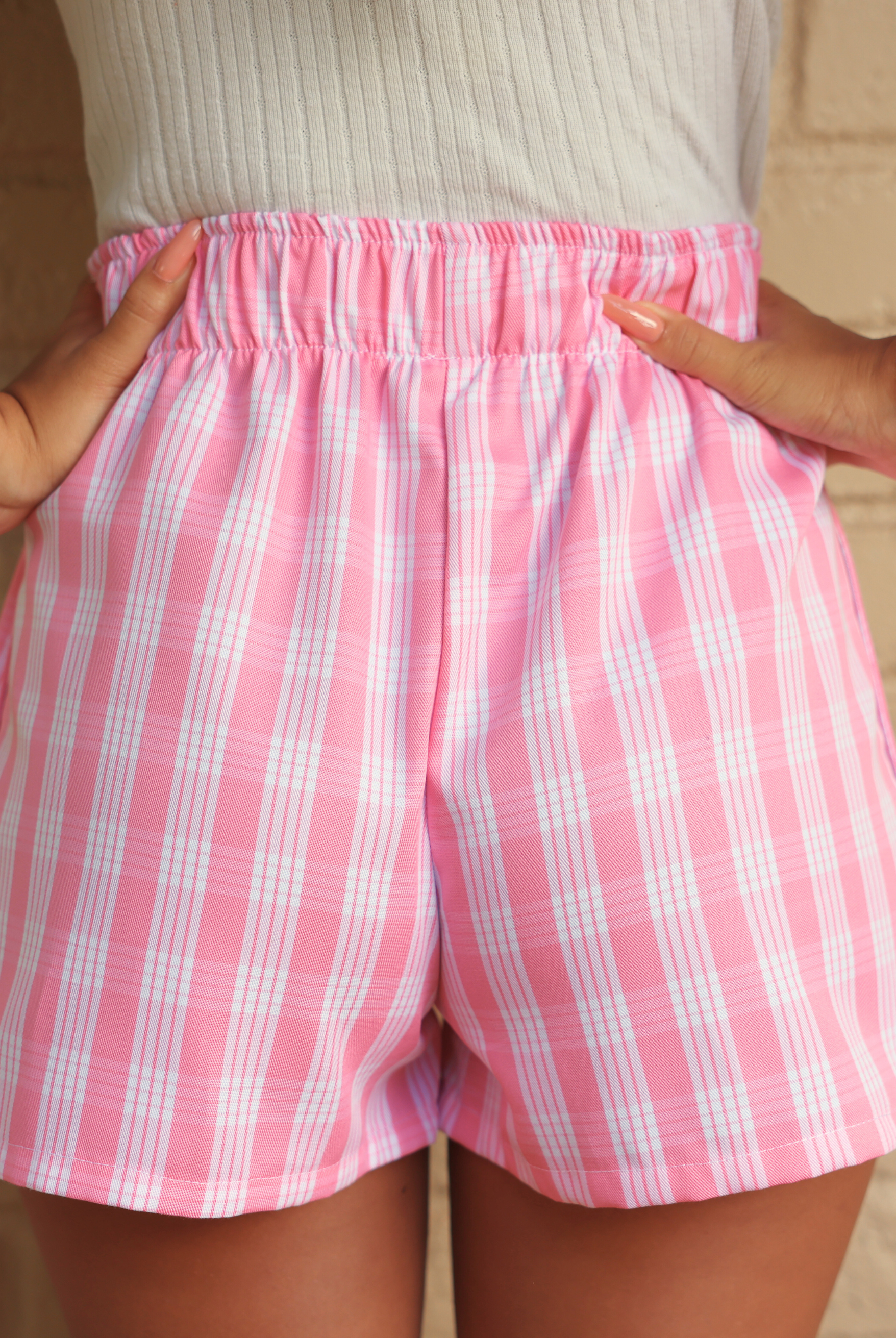 Wahine Holoholo Shorts 2.0 in Lokelani Light, featuring a pink Palaka pattern. Model poses with hands in pockets, showcasing the elastic waistband against a neutral background.