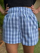 Wahine Holoholo Shorts 2.0 in Palaka print with elastic waist. Model's hands on hips, wearing a black top, outdoors in sunlight.