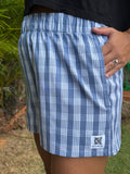 Wahine Holoholo Shorts 2.0 in traditional Palaka pattern, side view. Hand in pocket, subtle outdoor lighting, visible logo patch.