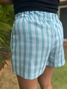 ʻEhukai shorts featuring a teal Palaka print, elastic waistband, and side view. Worn outdoors with greenery background.