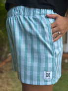 Wahine Holoholo Shorts in ʻEhukai tone with traditional Palaka print. Close-up side view shows elastic waistband and logo patch. Hand in pocket, wearing rings.