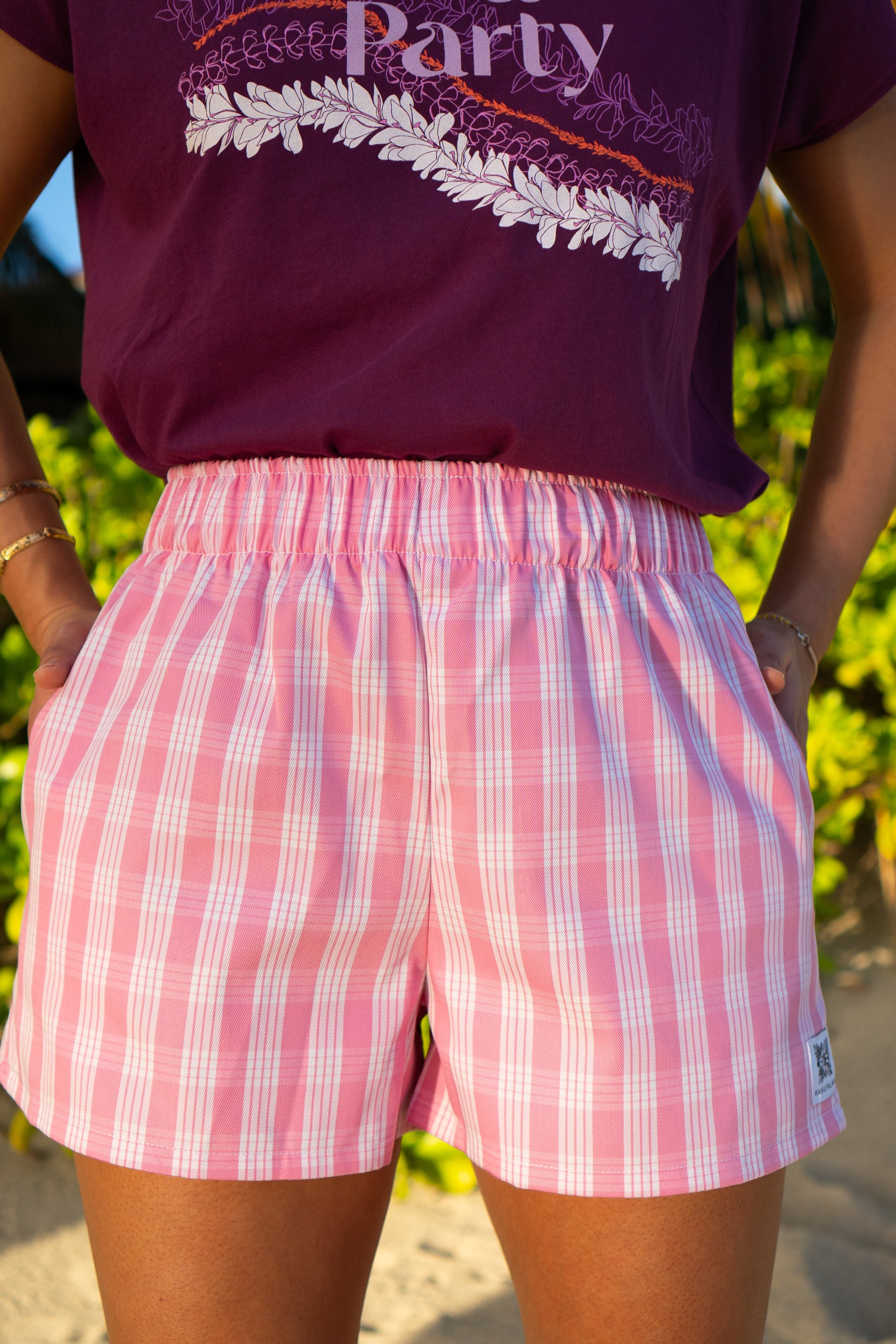 Pink Wahine Holoholo shorts in Palaka print, paired with a purple top. Bright outdoor lighting highlights the elastic waistband.