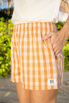 Wahine Holoholo Shorts 2.0 in Kaunaʻoa Rust Palaka print, with one hand in pocket. Bright outdoor lighting, greenery background.