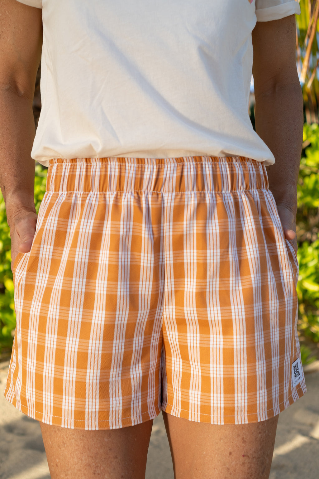Wahine Holoholo Shorts 2.0 in Kaunaʻoa Rust feature a Palaka print. The model poses with hands in pockets, wearing a white top. Bright sunlight enhances the orange tone.