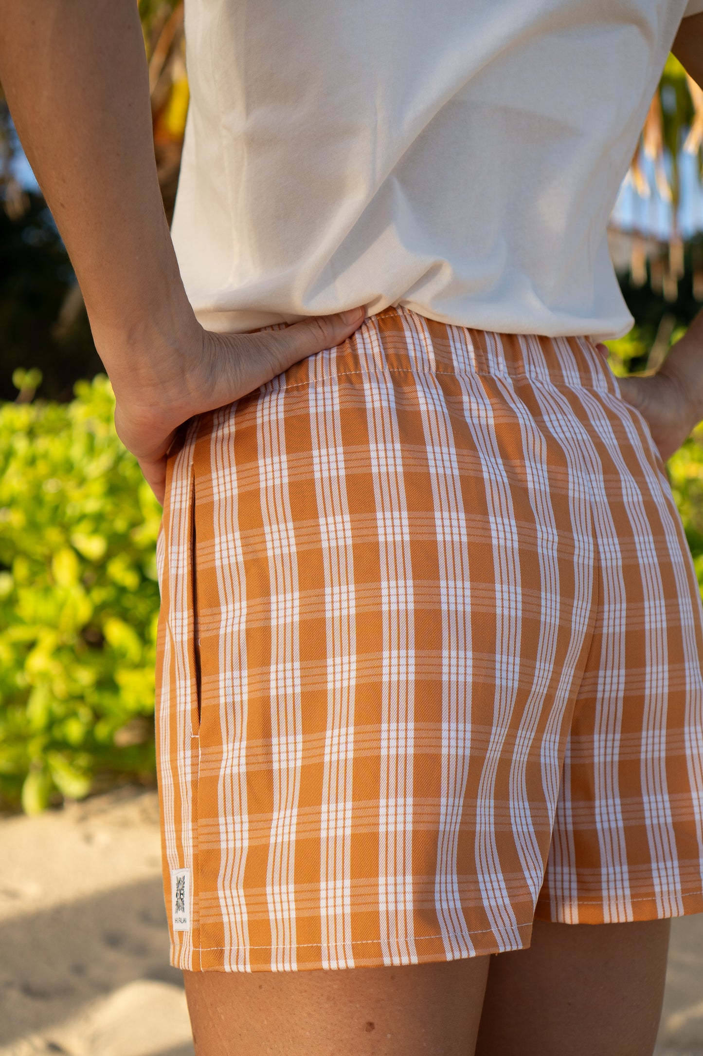 Wahine Holoholo shorts in Kaunaʻoa Rust with Palaka print. Model has hand on hip, wearing a white top. Bright outdoor lighting.