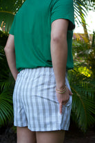 Wahine Holoholo Shorts 2.0 feature a Palaka print. Model poses with hand in pocket, wearing a green shirt and gold bracelets, surrounded by foliage.