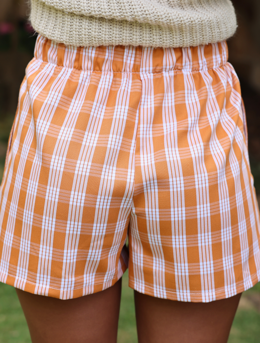 Wahine Holoholo Shorts 2.0 in Kaunaʻoa Rust Palaka print. Elastic waistband, paired with a beige knit top, outdoor setting.