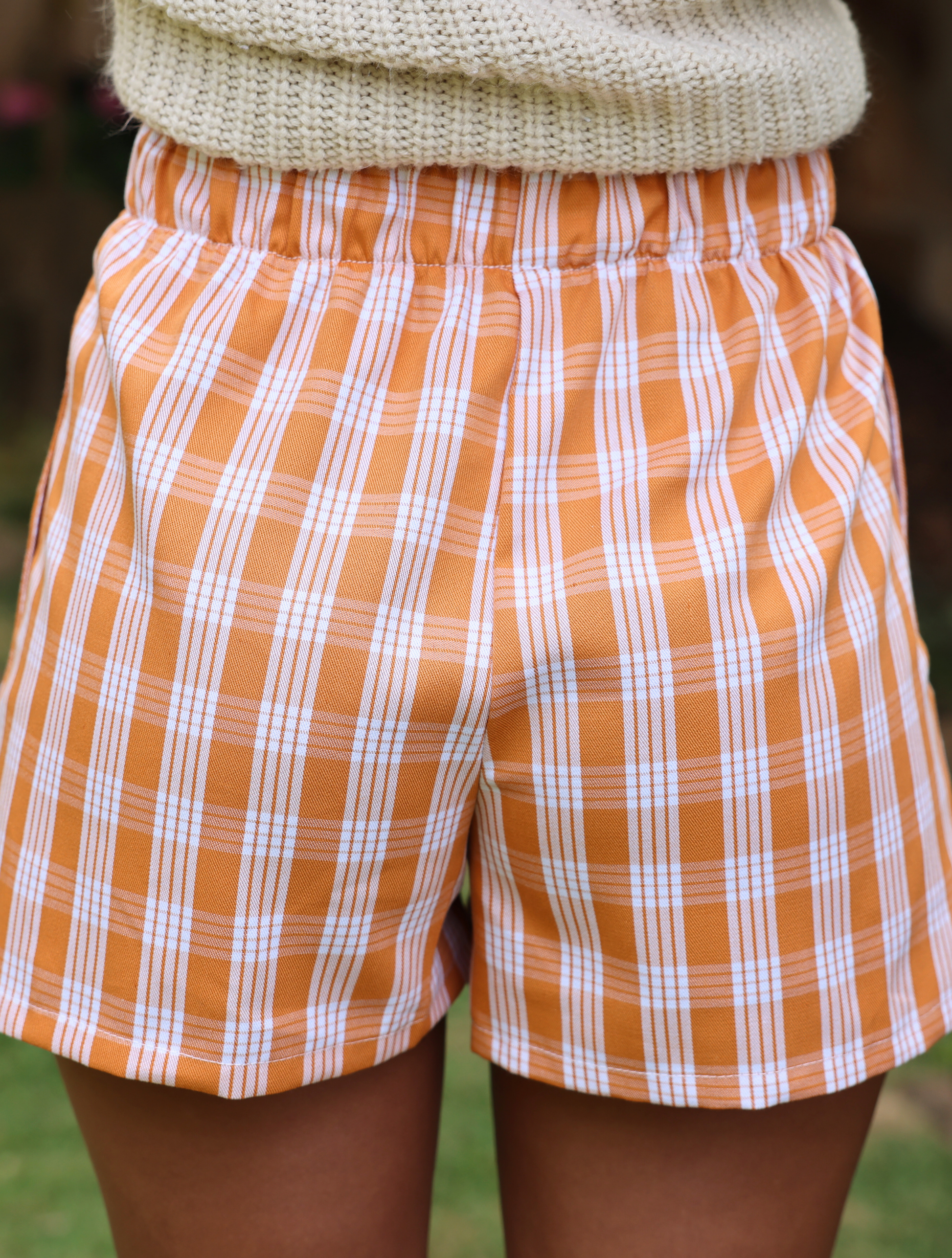 Wahine Holoholo Shorts 2.0 in Kaunaʻoa Rust Palaka print. Elastic waistband, paired with a beige knit top, outdoor setting.