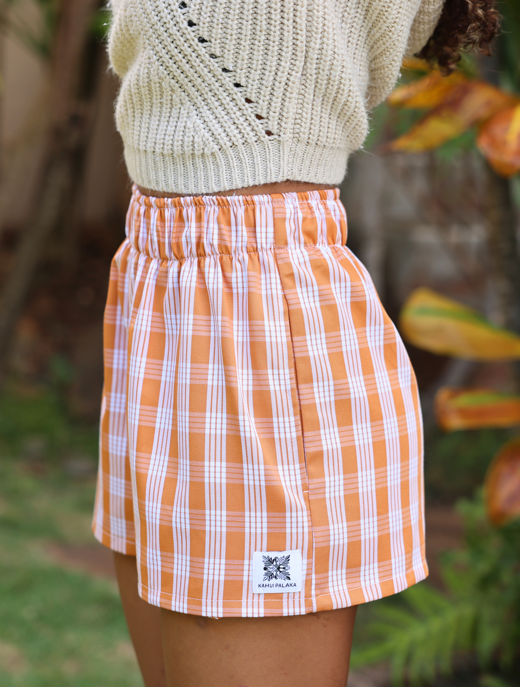 Wahine Holoholo Shorts 2.0 in Kaunaʻoa Rust with a traditional Palaka pattern. Elastic waistband, paired with a cream knit sweater.