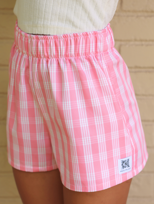 Wahine Holoholo Shorts 2.0 in Lokelani Light, featuring a pink Palaka pattern. Elastic waistband, side view, worn with a ribbed beige top.