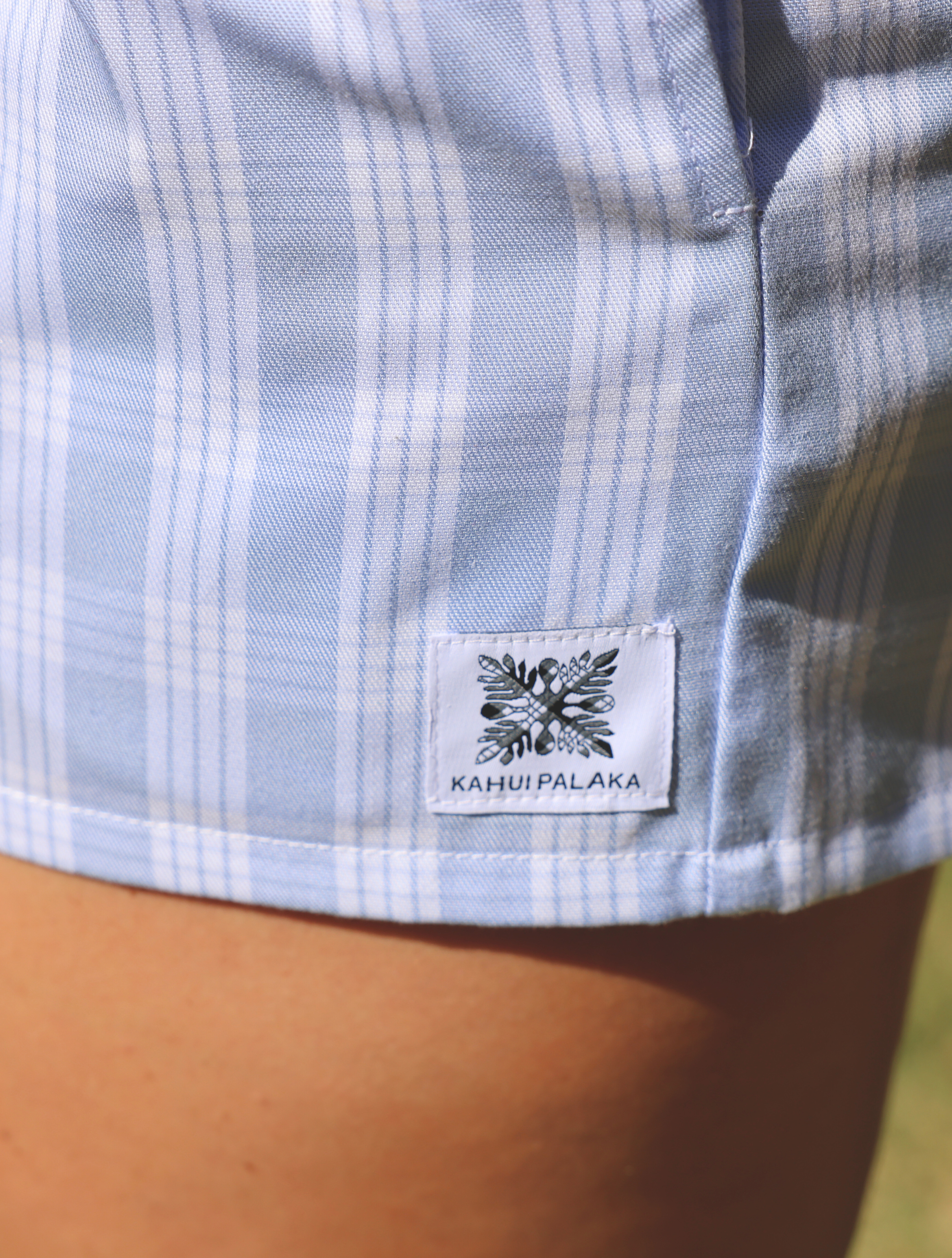 Wahine Holoholo Shorts 2.0 in light blue Palaka pattern. Close-up of side seam with brand tag. Sunlit, natural setting.