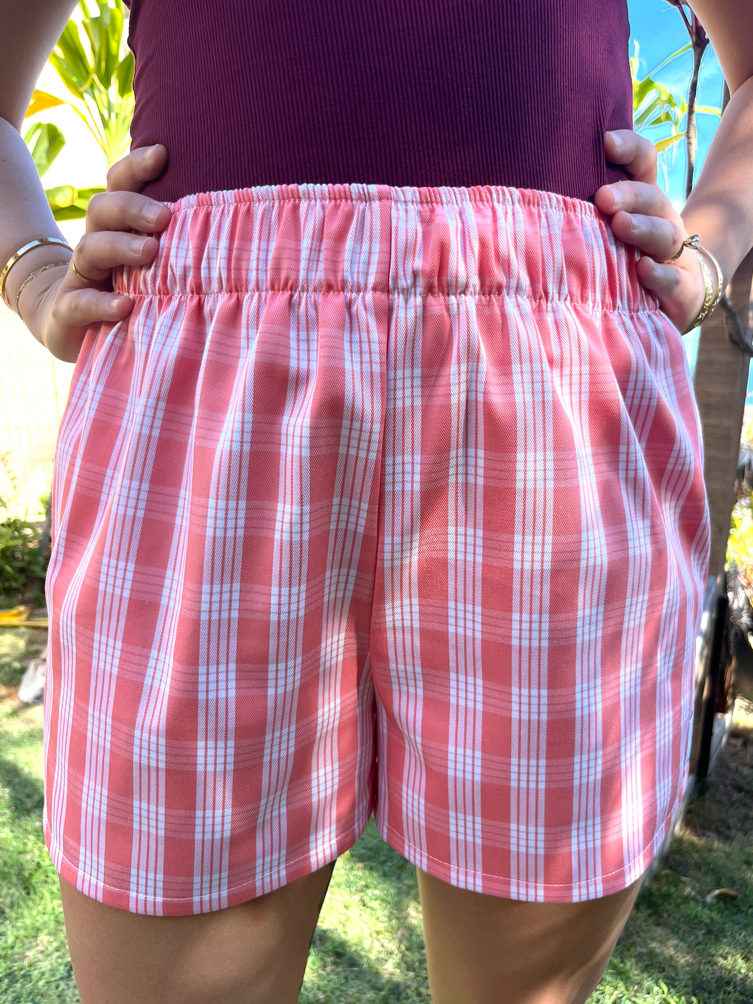 Wahine Holoholo Shorts 2.0 in ʻŌhelohelo feature a traditional Palaka print. Model poses with hands on hips. Bright sunlight highlights the shorts' pattern and colors.