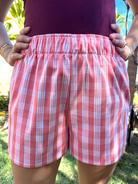Wahine Holoholo Shorts 2.0 in ʻŌhelohelo feature a traditional Palaka print. Model poses with hands on hips. Bright sunlight highlights the shorts' pattern and colors.