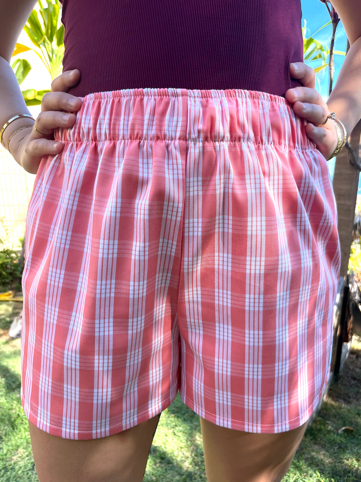 Wahine Holoholo Shorts 2.0 in ʻŌhelohelo feature a traditional Palaka print. Model poses with hands on hips. Bright sunlight highlights the shorts' pattern and colors.